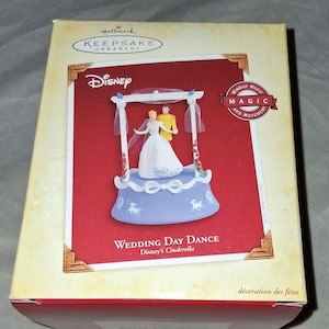 May include: A Hallmark Keepsake ornament featuring Disney's Cinderella. The ornament depicts Cinderella and Prince Charming dancing on a blue base with white accents. The box is tan with red and gold details, and the text "Wedding Day Dance" is visible.