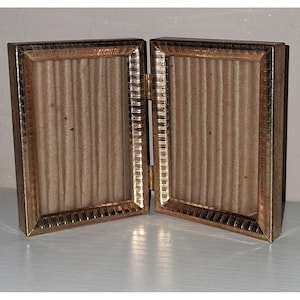 May include: A hinged, gold-colored metal picture frame, open to display two photo spaces. The frame has a textured border and a beige, vertically ribbed backing. The hinge is visible, connecting the two sides.