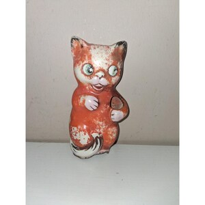 May include: A vintage-style cat figurine featuring a distressed orange and white paint finish. The cat has large green eyes, a small open mouth, and a curled tail. The surface shows signs of wear with chipped paint, revealing the base material.