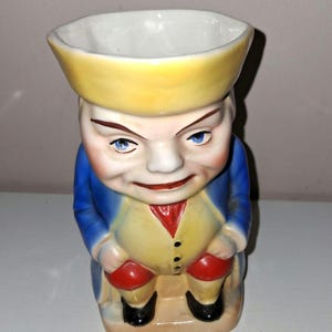 May include: A vintage ceramic figurine in the shape of a man with a stern expression. The figure wears a yellow hat, blue jacket, yellow vest, red breeches, and black shoes. The mug has a white interior.