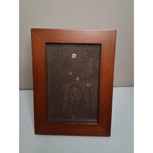May include: A rectangular wooden picture frame with a dark brown finish. The frame has a brown fabric backing with several brass-colored fasteners. The frame is empty and ready for a photo or artwork.