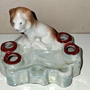 Pode incluir: Estatueta de cerâmica de um cão com marcas castanhas e brancas, pousada sobre uma base azul clara com quatro orifícios com bordas vermelhas. Esta peça vintage pode ser um castiçal ou um elemento decorativo.