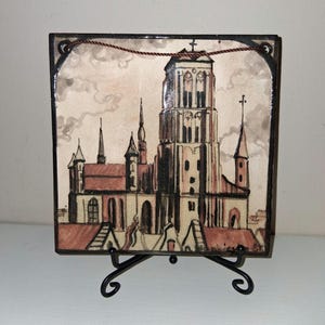 Puede incluir: Azulejo de cerámica cuadrado con una pintura de acuarela de una catedral con una torre alta y múltiples agujas. El azulejo se exhibe en un soporte de metal negro. La obra de arte tiene una paleta de colores apagados beige, marrón y rojo.