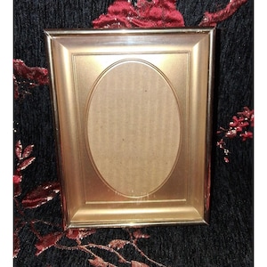 May include: A gold-colored picture frame with an oval opening. The frame has a rectangular shape with a slightly raised border. The background is a dark fabric with red floral accents. The frame is designed to hold a photograph or artwork.
