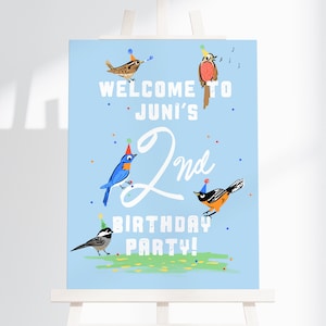 May include: A light blue sign with the text "WELCOME TO JUNI'S 2nd BIRTHDAY PARTY!" is decorated with illustrations of birds wearing party hats. The sign is displayed on a white easel.