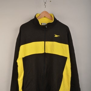 Op de afbeelding: Zwart en geel jack met rits en een Reebok-logo op de borst.