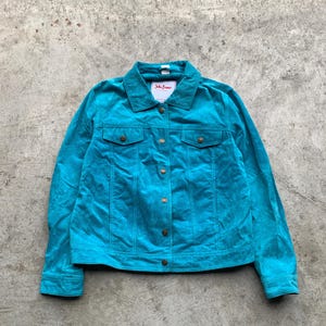 May include: A turquoise suede jacket with a collared neckline, two chest pockets, and button closures. The jacket has a label that reads "John Banner". The jacket is laid flat on a concrete surface.