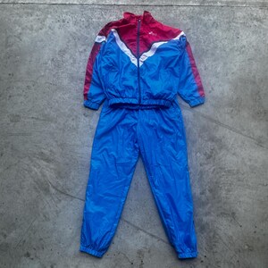 Op de afbeelding: Een vintage trainingspak in blauw en rood. De jas heeft een rood bovenstuk met witte en blauwe accenten, een ritssluiting en het merklogo. De broek is effen blauw. Het trainingspak is gemaakt van glanzend, lichtgewicht materiaal.