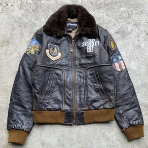 May include: A vintage brown leather bomber jacket with a brown fur collar. The jacket features multiple patches, a front zipper, and brown ribbed cuffs and hem. The word "Schott" is visible on the collar and chest.