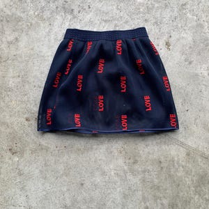 Op de afbeelding: Een marineblauwe minirok met het woord "LOVE" in het rood gedrukt. De rok heeft een geplooid ontwerp en een comfortabele elastische tailleband. Het woord "LOVE" wordt meerdere keren herhaald over de stof.