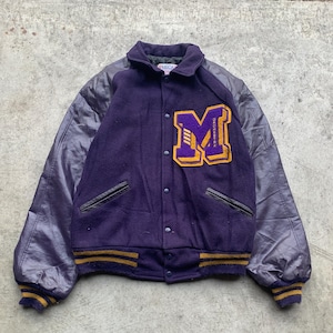 Könnte beinhalten: Eine violette Vintage-Collegejacke mit einem großen gelb-violetten "M"-Aufnäher auf der Brust. Die Jacke hat dunkelviolette Lederärmel, eine Knopfleiste und gelb-violett gestreifte Bündchen und einen Bund.