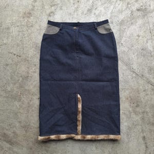 Könnte beinhalten: Dunkelblauer Jeansrock mit hoher Taille und Frontschlitz. Der Rock hat graue Akzente an den Taschen und einen Kunstfellbesatz am unteren Rand. Der Rock hat eine Midi-Länge.