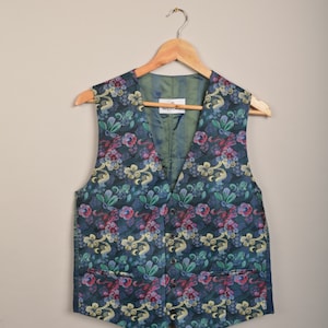 Op de afbeelding: Een donkerblauw vest met een bloemenpatroon in tinten roze, geel en groen. Het vest heeft een V-hals en is gemaakt van een zijdeachtige stof. Het label op het vest luidt "Wilvorst".