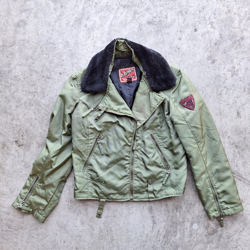 K&ouml;nnte beinhalten: Olivgr&uuml;ne Motorradjacke mit schwarzem Kunstfellkragen. Die Jacke hat mehrere Rei&szlig;verschlusstaschen, einen roten Aufn&auml;her am &Auml;rmel und den Markennamen "h.brides" auf einem Etikett. Die Jacke ist aus gl&auml;nzendem, wasserabweisendem Material.