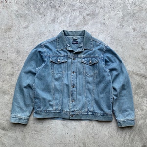 Op de afbeelding: Lichtblauwe spijkerjas met een knoopsluiting, twee borstzakken en een klassieke kraag. De jas heeft een licht vervaagde uitstraling, wat wijst op een vintage stijl. De merknaam is zichtbaar op de kraag.