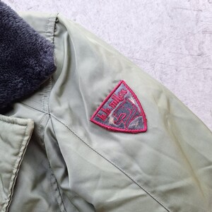 K&ouml;nnte beinhalten: Nahaufnahme einer olivgr&uuml;nen Jacke mit einem dunkelgrauen Kunstfellkragen. Ein roter und grauer Aufn&auml;her auf dem &Auml;rmel zeigt den Text "kk miles" und eine Pferde-Grafik. Die Jacke hat einen Knopfverschluss.