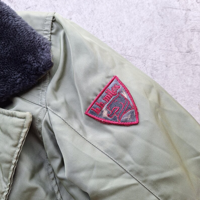 K&ouml;nnte beinhalten: Nahaufnahme einer olivgr&uuml;nen Jacke mit einem dunkelgrauen Kunstfellkragen. Ein roter und grauer Aufn&auml;her auf dem &Auml;rmel zeigt den Text "kk miles" und eine Pferde-Grafik. Die Jacke hat einen Knopfverschluss.
