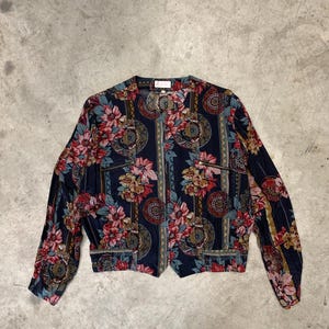 Op de afbeelding: Donkerblauwe jas met bloemenprint, ronde hals en lange mouwen. De jas heeft een levendig patroon van rode, roze en gele bloemen, met decoratieve cirkelvormige en gestreepte accenten. De jas heeft twee voorzakken en een ritssluiting.