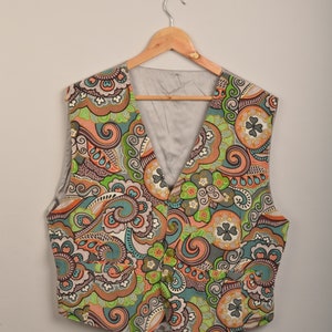 Op de afbeelding: Een vest met een paisley-patroon in tinten groen, oranje, bruin en wit. Het vest hangt aan een houten hanger.