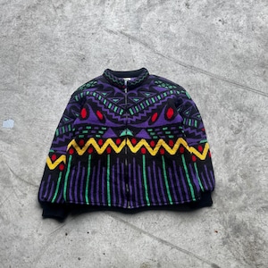 Op de afbeelding: Een vintage jas met ritssluiting en een levendig geometrisch patroon. De jas heeft een zwarte basis met paarse, groene, gele en rode accenten. De kraag en manchetten zijn effen zwart.