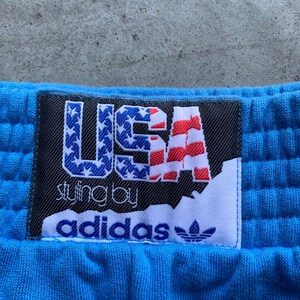 Może przedstawiać: Niebieskie spodnie dresowe z czarną i białą metką z napisem "USA styling by adidas" i wzorem amerykańskiej flagi w kolorze czerwonym, białym i niebieskim.