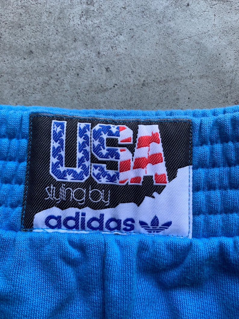 Może przedstawiać: Niebieskie spodnie dresowe z czarną i białą metką z napisem "USA styling by adidas" i wzorem amerykańskiej flagi w kolorze czerwonym, białym i niebieskim.