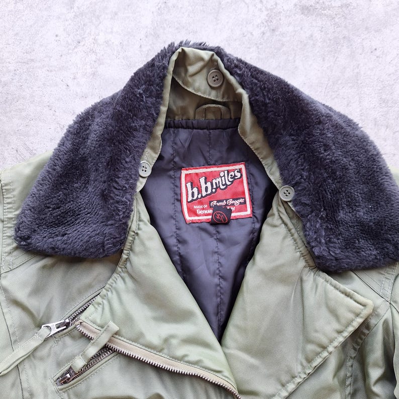K&ouml;nnte beinhalten: Nahaufnahme einer olivgr&uuml;nen Jacke mit einem dunkelgrauen Kunstfellkragen. Die Jacke hat einen Rei&szlig;verschluss und ein rotes Etikett mit der Aufschrift "b.b.miles". Der Kragen hat zwei Kn&ouml;pfe.