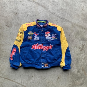 Op de afbeelding: Blauwe en gele racejas met diverse sponsorlogo's, waaronder "Kellogg's" in rode letters. De jas heeft een knoopsluiting en een stervormig embleem aan de onderkant.