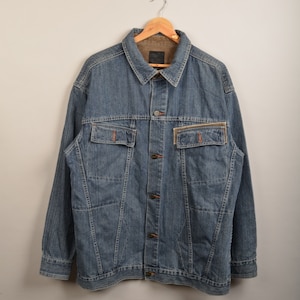 Op de afbeelding: Een blauwe denimjas met een bruine corduroykraag en een ritszak aan de linkerkant. De jas heeft een knoopsluiting en meerdere zakken.