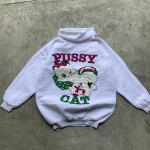 Op de afbeelding: Witte coltrui met de woorden "PUSSY CAT" in roze en groen. De afbeelding toont een cartoonkat met een roze strik en een groen-wit stippenpatroon.