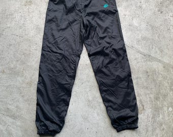 Könnte beinhalten: Schwarze Nylon-Trainingshose mit elastischem Bund und Knöchelbündchen. Ein kleines türkisfarbenes Nike-Logo ist auf dem linken Bein zu sehen. Wahrscheinlich im Vintage- oder Retro-Stil.