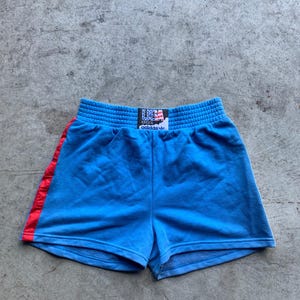 Może przedstawiać: Niebieskie spodenki sportowe z czerwonym paskiem po boku i czarnym pasem w talii z napisem "USA" i "adidas" w kolorze białym.