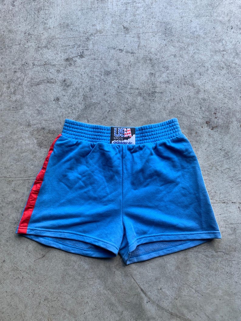 Może przedstawiać: Niebieskie spodenki sportowe z czerwonym paskiem po boku i czarnym pasem w talii z napisem "USA" i "adidas" w kolorze białym.