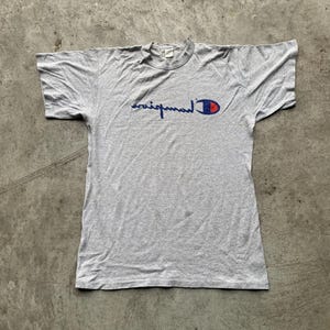 Op de afbeelding: Een grijs Champion T-shirt met een ronde hals en korte mouwen. Het Champion-logo staat in blauw schrift met een rood-blauw embleem op de linkerborst. Het shirt ligt plat op een grijze ondergrond.