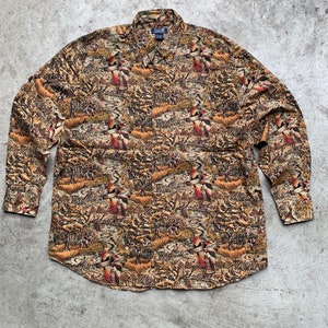 May include: A long-sleeved button-down shirt with a brown, green, and orange camouflage pattern featuring birds. The shirt has a button-down collar and a single chest pocket.