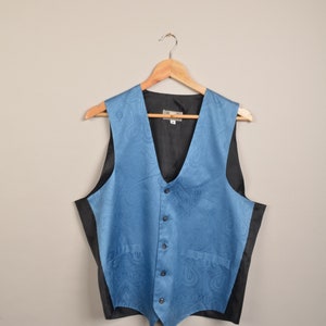 Puede incluir: Un chaleco azul claro con estampado de cachemira y espalda y forro negros, colgado de una percha de madera. El chaleco tiene cuello en V y cierre de botones. El estampado de cachemira es visible en la parte delantera.