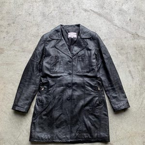 Op de afbeelding: Een zwarte leren trenchcoat met een knoopsluiting en twee ritszakken aan de zijkanten.