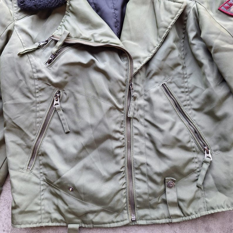 K&ouml;nnte beinhalten: Olivgr&uuml;ne Jacke mit mehreren Rei&szlig;verschlusstaschen und einem diagonalen Rei&szlig;verschluss. Die Jacke hat einen schwarzen Kunstfellkragen und einen kleinen roten Aufn&auml;her auf dem rechten oberen &Auml;rmel. Die Jacke hat ein gestepptes Futter.