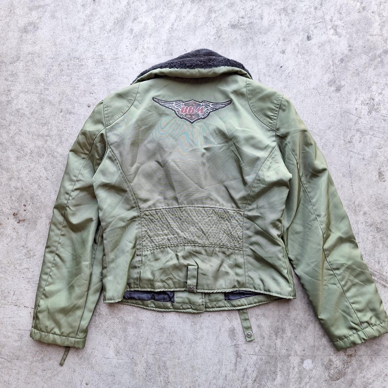 K&ouml;nnte beinhalten: Olivgr&uuml;ne Jacke mit schwarzem Kragen und Harley-Davidson-Logo auf dem R&uuml;cken. Die Jacke hat ein gestepptes Design am unteren R&uuml;cken und einen Knopfverschluss. Die Jacke ist aus leichtem Material gefertigt.