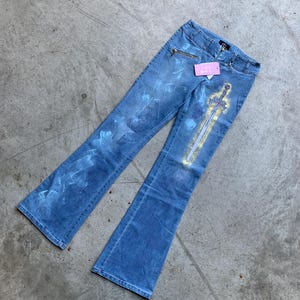 May include: Light blue denim flare jeans featuring a faded white wash pattern and a prominent yellow and white sword graphic printed on the left leg. The jeans include a zippered pocket on the right hip, a button closure, and a small pink tag.