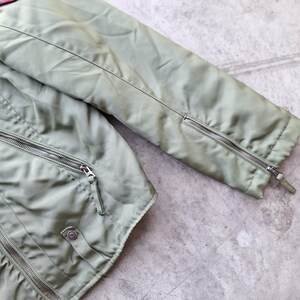 K&ouml;nnte beinhalten: Nahaufnahme einer salbeigr&uuml;nen Jacke mit silbernen Rei&szlig;verschl&uuml;ssen. Die Jacke hat einen Rei&szlig;verschluss, einen Druckknopf und Rei&szlig;verschl&uuml;sse an den &Auml;rmeln. Der Stoff hat einen leichten Glanz und die Jacke scheint aus einem leichten Material zu sein.