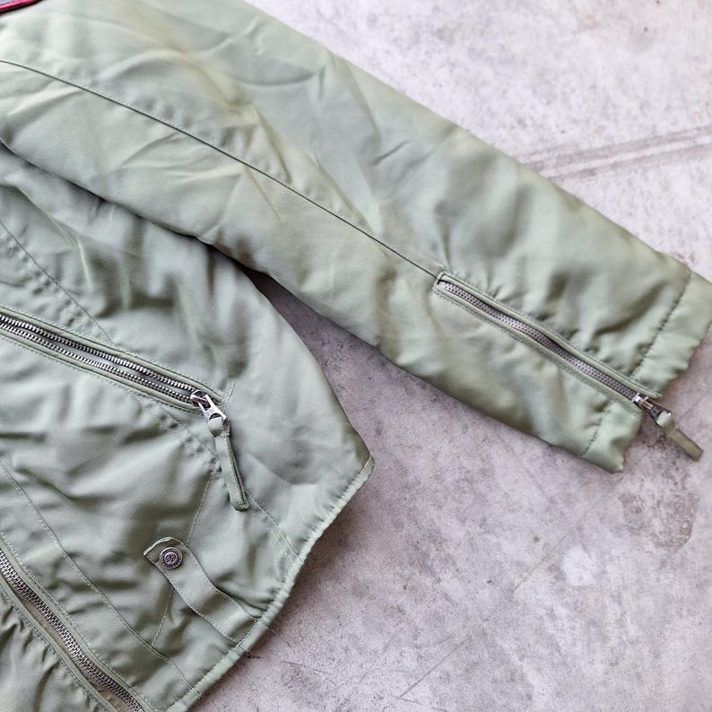 K&ouml;nnte beinhalten: Nahaufnahme einer salbeigr&uuml;nen Jacke mit silbernen Rei&szlig;verschl&uuml;ssen. Die Jacke hat einen Rei&szlig;verschluss, einen Druckknopf und Rei&szlig;verschl&uuml;sse an den &Auml;rmeln. Der Stoff hat einen leichten Glanz und die Jacke scheint aus einem leichten Material zu sein.
