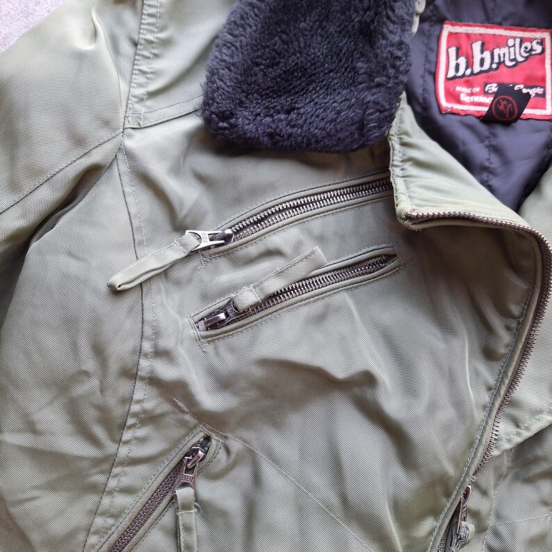 K&ouml;nnte beinhalten: Nahaufnahme einer olivgr&uuml;nen Jacke mit mehreren silbernen Rei&szlig;verschl&uuml;ssen und einem dunkelgrauen Kunstfellkragen. Die Jacke hat ein Etikett mit der Aufschrift "b.b.miles". Die Jacke ist aus einem glatten, m&ouml;glicherweise Nylon-Material.
