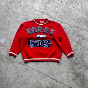 Op de afbeelding: Een rode trui met de tekst "AVIREX" in grijs en "CADET" in blauw. De trui heeft witte strepen op de manchetten en zoom. De trui is gemaakt van gebreid materiaal.