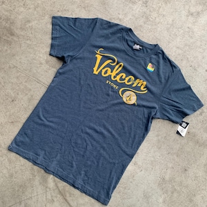 May include: A blue short-sleeve t-shirt with the word "Volcom" in yellow script across the chest. Below the word "Volcom" is the word "STONE" in a smaller font. A small graphic is below the word "STONE". A tag is attached to the shirt.