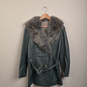 Op de afbeelding: Een tealblauwe leren jas met een grote grijze nep bontkraag en een bijpassende riem. De jas heeft een dubbele rij knopen en twee voorzakken.