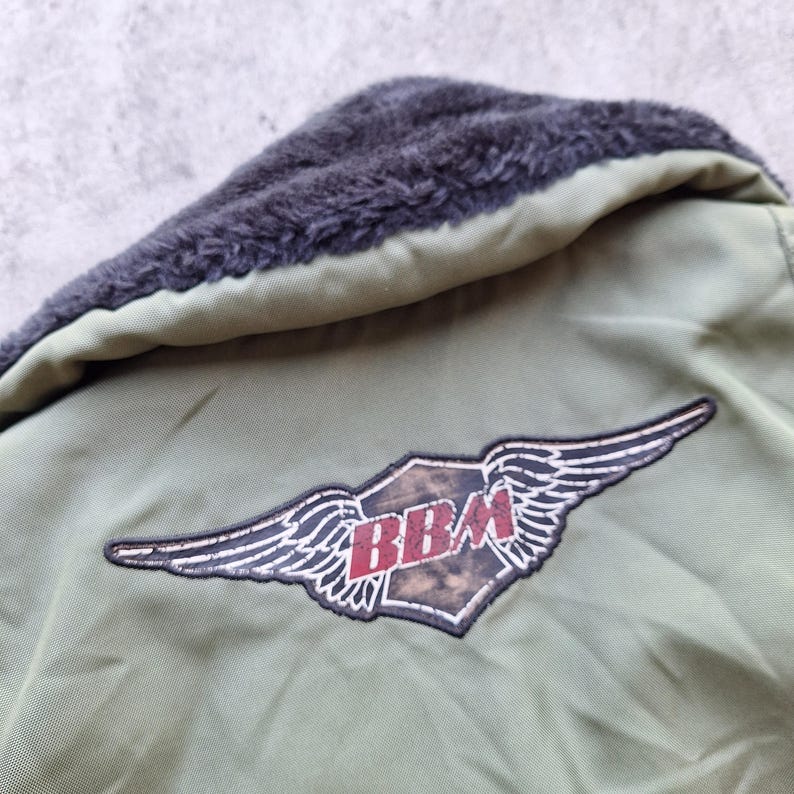 K&ouml;nnte beinhalten: Nahaufnahme einer olivgr&uuml;nen Jacke mit einem dunkelgrauen Kunstfellkragen. Die Jacke hat einen Aufn&auml;her mit den roten Buchstaben "BBM" in einem gefl&uuml;gelten Emblem.