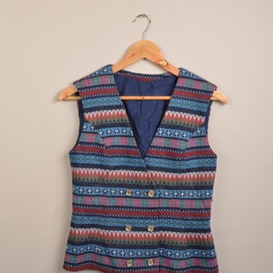 Puede incluir: Un chaleco sin mangas con cuello en V y un colorido estampado geométrico en azul, rojo y verde. El chaleco tiene un diseño de doble botonadura con botones dorados y está colgado de una percha de madera.