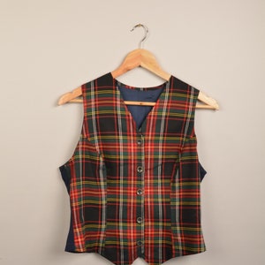 Op de afbeelding: Een vest zonder mouwen met een geruit patroon in rood, groen, zwart en geel. Het vest heeft een V-hals en wordt gesloten met knopen.