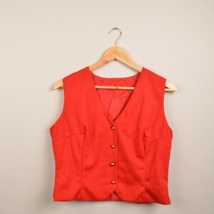 Op de afbeelding: Een rood vest zonder mouwen met gouden knopen hangend aan een houten hanger tegen een witte achtergrond.